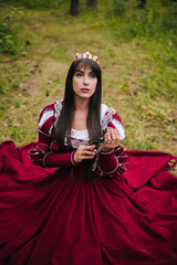 Fototapeta premium A beautiful girl with black long hair in a medieval, red dress with a crown on her head walks in the forest. Fairy-tale princess. Fantasy