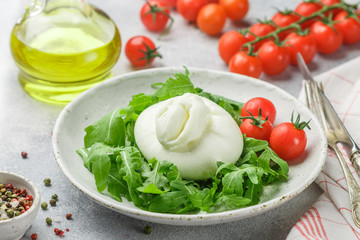 Delicious Italian fresh burrata cheese with arugula salad, cherry tomatoes and olive oil in a white plate. Gourmet snack