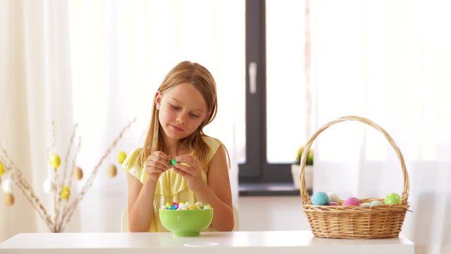 easter, holidays and people concept - happy girl removing wrapper from chocolate egg candy at home