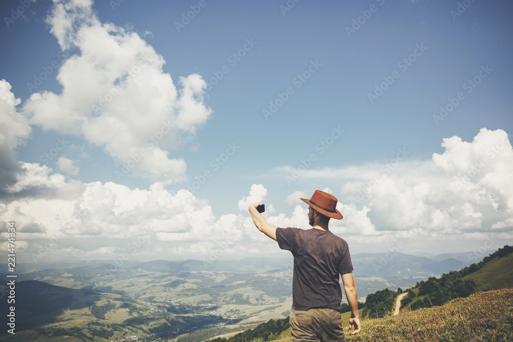 Wall mural stylish traveler man in hat holding phone, walking on top of sunny mountains and sky. travel and wanderlust concept. space for text. happy hipster traveling and taking photo.