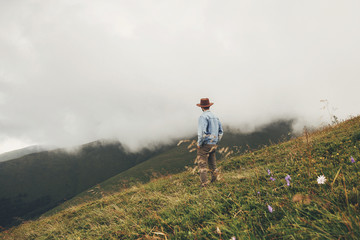 travel and wanderlust concept. stylish traveler man in hat walking on top of  mountains in grass into the clouds. space for text. hipster guy traveling. amazing atmospheric moment