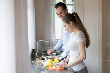 Obraz na płótnie Canvas Happy young parents prepare healthy breakfast at home together, smiling husband and wife making food in new house kitchen, millennial man and woman spend weekend morning cooking delicious food