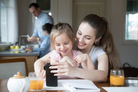Happy mother and daughter having fun playing game on smartphone in kitchen, mom and girl spending time together watching cartoons or children video on phone, parent teach little kid using cell
