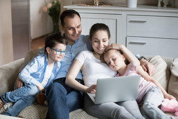 Smiling young family lying with kids on cozy sofa watching cartoons on laptop, happy parents hug...