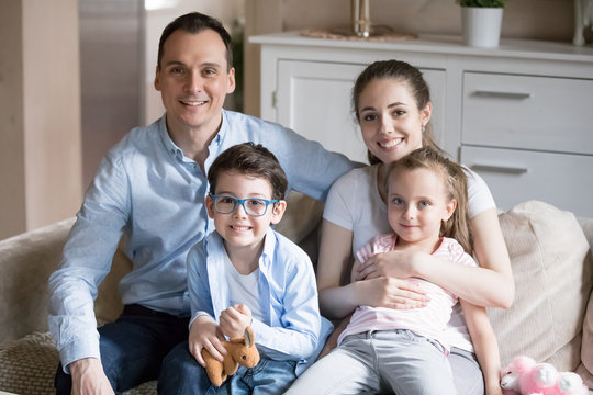 Portrait of happy family of four sit on couch in living room, smiling young parents hug small kids taking picture together, mom and dad embrace son and daughter when making photo for album in house