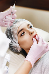 Preparation process for the lip enhancement. The doctor draws up  hyaluronic acid in front of the patient. The young girl with a beautiful face in the special hat and hands of the doctor in pink glove