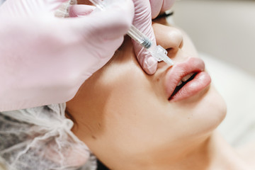 Obraz na płótnie Canvas The process of lips enhancement. Cosmetician makes an injection of hyaluronic acid in a beautiful lips. The young girl with a beautiful face in the special hat and hands of the doctor in the pink glov