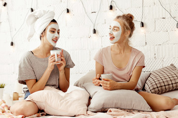 Portrait of beautiful smiling girls with cosmetic cleansing masks sitting in bed drinking hot...