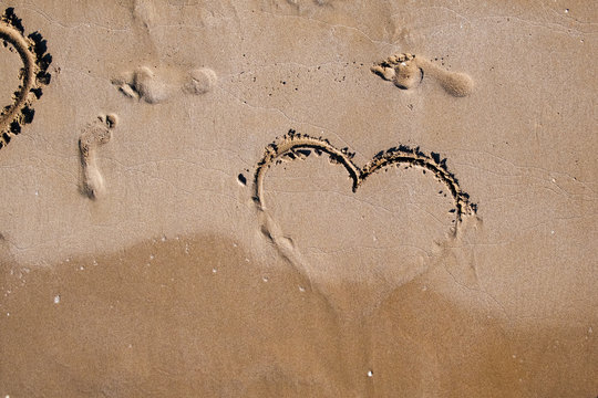 emblem of the heart of love is painted on the sea sand on a warm summer day next to the prints of human feet