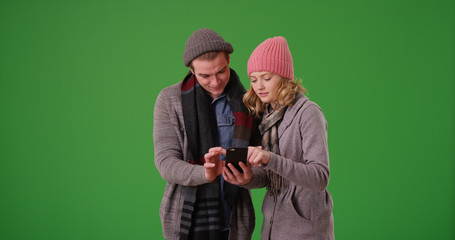 Young white couple in winter attire sharing smartphone on green screen