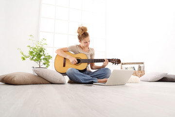 Fototapeta premium Piękna młoda kobieta gra na gitarze z komputerem, uczy się grać na kursie online, siedząc na podłodze w zwykłym ubraniu w domu