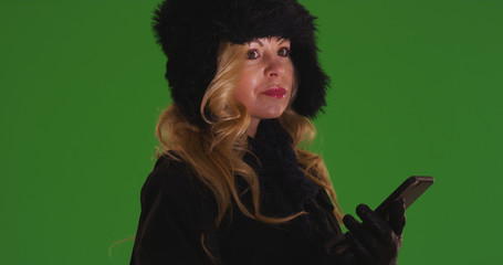 Caucasian woman in winter attire with cellphone looking around on green screen