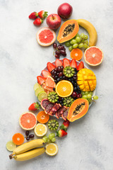 Raw fruits berries platter, mango, oranges, kiwi strawberries, blueberries grapefruit grapes, bananas apples on the white plate, on the off white table, top view, vertical, copy space