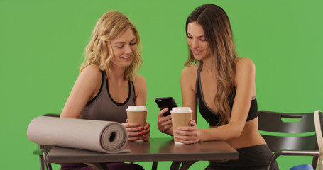 Obraz na płótnie Canvas Two healthy yoga girls sitting at cafe sharing smartphone on green screen