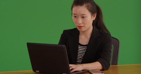 Asian businesswoman at workstation typing on laptop on green screen