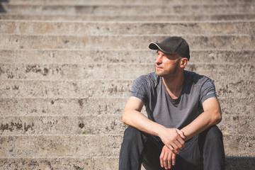 a man with a stubble in a baseball cap sits resting on the steps and enjoying the sun, toned