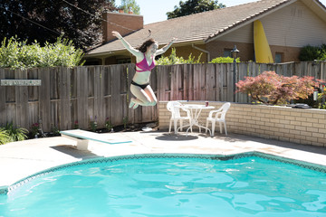 Young woman jumping off diving board into a backyard swimming pool arms up in the air legs tucked to her body smiling caught in mid air. Girl in bikini jumps into outdoor swimming pool in a backyard.