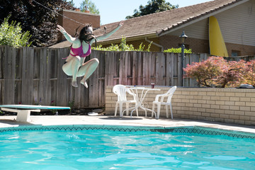 Young woman jumping off diving board into a backyard swimming pool arms up in the air legs tucked to her body caught in mid air. Girl in bikini jumps into outdoor swimming pool in the backyard.