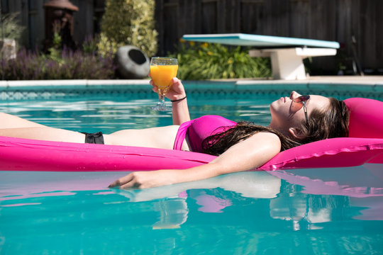 Woman lounging in an outdoor backyard swimming pool on a pink raft holding a drink in her hand on a sunny summer day. Diving board and garden in the background. Relaxing in a pool.