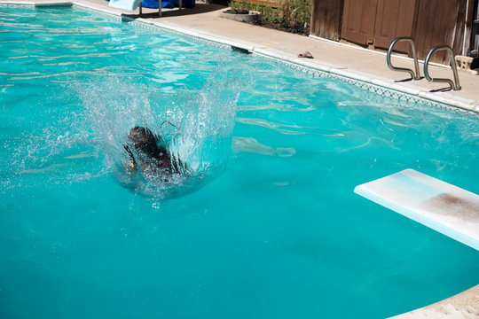 Dark haired woman doing a cannon ball into an outdoor swimming pool in a sunny summer day. Young woman jumps off diving board into a backyard swimming pool in the summer.