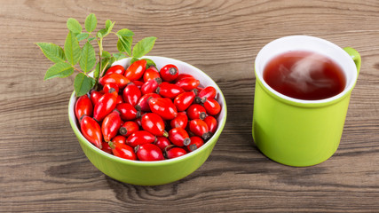 Rosehip tea and red briar fruit pile on brown wood background. Rosa canina. Ceramic mug with smoking medicinal drink. Beautiful bowl full of fresh ripe wild hips and decorative twig with green leaves.