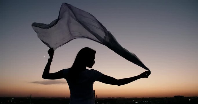 Silhouette of Young pretty girl standing on the roof, holding up her fabric in wind closeup 4k
