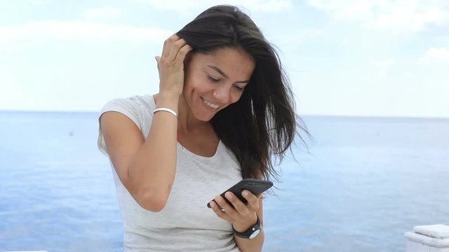 Smiling young woman satisfied with received news via her smartphone while enjoying the sea. Happiness, joyous moments, feeling good. Positive mood. Female portrait
