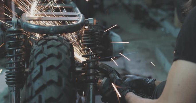 Man works circular saw. Flies of spark from hot metal. Handsome guy working with custom motorcycle in the garage. Close-up of hand and electric saws metal hand tool