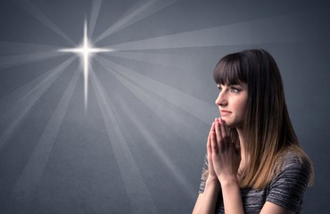 Young woman praying on a grey background with a shiny cross silhouette above her