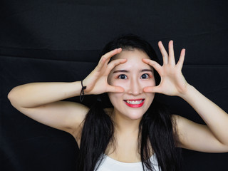 Fototapeta premium Portrait of good looking sexy Chinese woman with long black hair and white halter against black background, smiling at camera with hands around eyes.