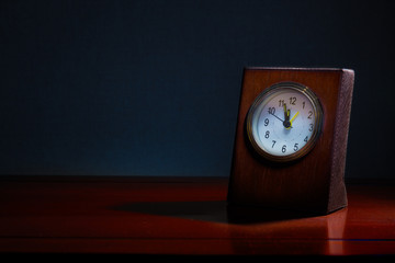 Old business desk clock on wooden table at night with the time showing almost midnight, blue tone background, good for time related concept