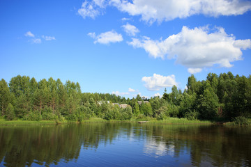Lake reflecting sky with clouds on it flowing through a small village and covering banks with trees, shrubs and plants growing on them