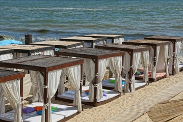 a row of brown open pavilions with white curtains on the beach sand near the sea
