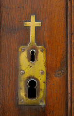 Copper or brass ornate plate with double keyhole, decorated with a cross on the wooden surface of church door