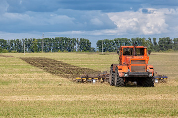 Obraz premium An orange modern tractor plows the earth in.&nbsp; a golden field of wheat on a summer day, in the sky a cumulus cloud, in the background a forest.