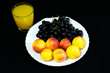 Peaches, black seedless grapes and nectarines. Orange juice and fruit for Breakfast. Yellow tropical fruit on a white plate. A glass of juice on a black background. Vegetarian and raw food menu. Diet