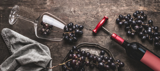 Red wine still life with bottle and vintage corkscrew, glass and grapes on aged wooden background,...