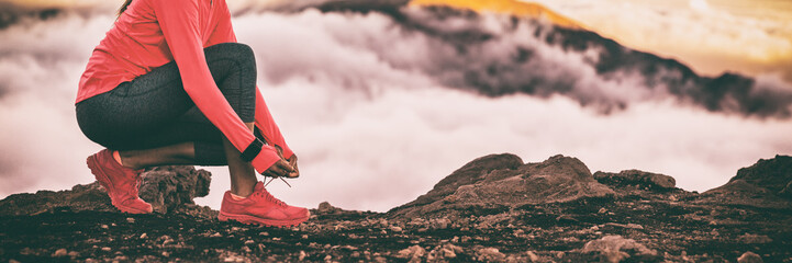 Runner smartwatch woman getting ready for trail run in high cold clouds mountains background tying up running shoes laces. Fitness and sports motivation healthy lifestyle. Banner panoramic crop.