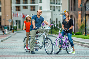 Fototapeta premium Happy Caucasian family of father, mother and daughter posing for camera outdoors in the street at summer on buildings background during bicycle ride. Active leisure and sports activities at resort