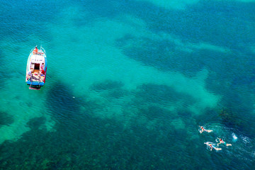 Aerial shot of turquoise water lagoon at hot summer day with white boat. Top view of people swimming around the boat.
