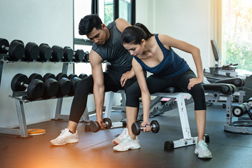 Fototapeta na wymiar The beauty lady and handsome man are wearing exercise suit,prepare for lifting dumbbell up,for built arm muscle,at fitness room,blurry light around
