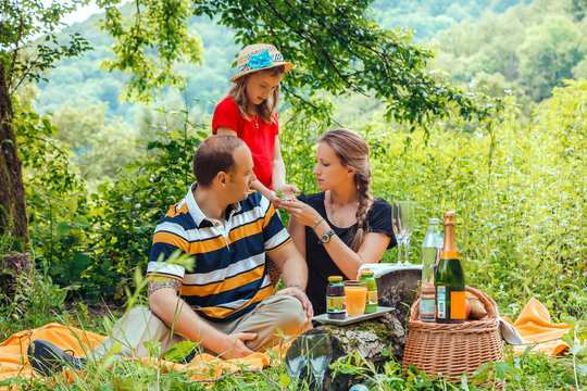 Young Happy Caucasian Family Of Father, Mother And Daughter Having Picnic On Vacation In Summer Mountain Forest