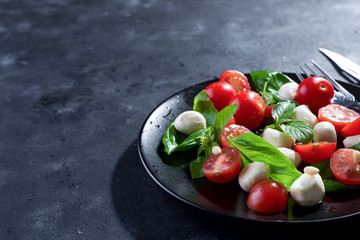 Italian caprese salad with sliced tomatoes, mozzarella cheese, basil, olive oil. Served in black plate with fork over dark metal background. Top view with space.