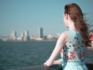 The young girl stands on the bridge and looks at the river. Summer travel trip. View from the back.