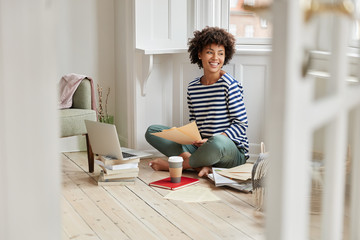 Cheerful smart female designer holds graphic illustration, works on new project, uses scientific literature, drinks takeaway coffee, feels carefree, enjoys domestic atmosphere, learns new information