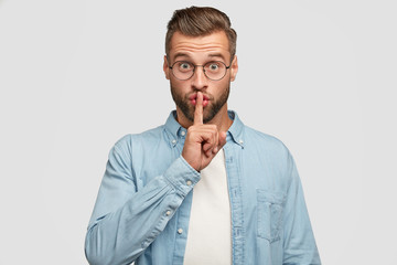 Shh, dont speak too loudly! Serious unshaven guy keeps index finger on lips, keeps conspiracy, asks not spread confidential information, dressed in casual outfit, isolated on white studio wall