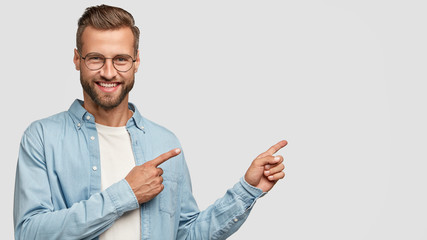 Positive unshaven man looks and points at upper right corner with both index fingers, smiles with...