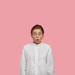 Vertical shot of terrified young woman with shocked expression, stares with unexpected gaze, opens mouth from wonder, wears casual white shirt, isolated over pink background. Girl in stupor.