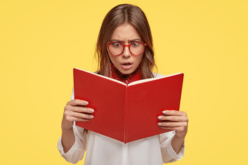 Photo of shocked beautiful student stares at opened red look, reads awful news, finds out unknown fact, stares through glasses, isolated over yellow studio wall. Teenager recieves knowledge.