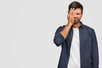 Indoor shot of handsome stressful overworked man covers face with palm, has displeased expression,...